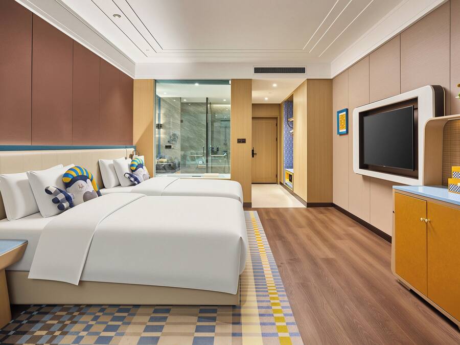 A hotel room with a bed, bedside tables, a TV, a minibar, and a bathroom visible through a glass door.