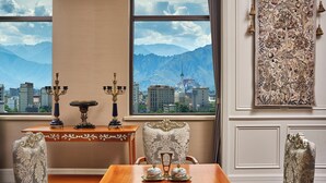 A room with a view of mountains and a cityscape through large windows, a table with decorative items, and a patterned wall hanging.