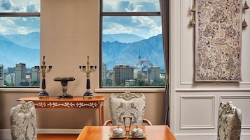 A room with a view of mountains and a cityscape through large windows, a table with decorative items, and a patterned wall hanging.