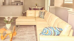 A living room with a beige sectional sofa, a glass coffee table, and a patterned rug. - Nyakio Suite - Chesapeake (Chesapeake)