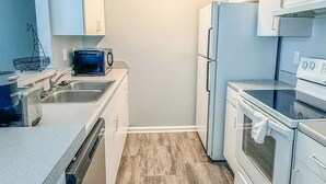 A compact kitchen with white cabinets, a built-in dishwasher, a microwave, a stove, and a refrigerator. - Nyakio Suite - Chesapeake (Chesapeake)