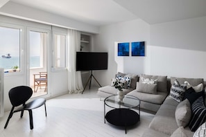 Ein modernes Wohnzimmer mit einer Couch, einem Couchtisch, einem Fernseher auf einem Schrank und Blick auf das Meer durch große Fenster.