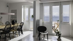 A dining area with a table and chairs, a radiator, and a view of the sea through large windows. - True Blue by halu! - Seafront 2BR apartment (Thessaloniki)
