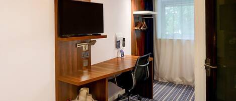 Une chambre d’hôtel avec un bureau en bois, une télévision, un porte-manteau et une fenêtre avec des rideaux.
