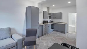 Een compacte keuken met grijze kastjes, een zithoek met een grijze bank en een zwarte stoel, en een vloer met een herringbone patroon.