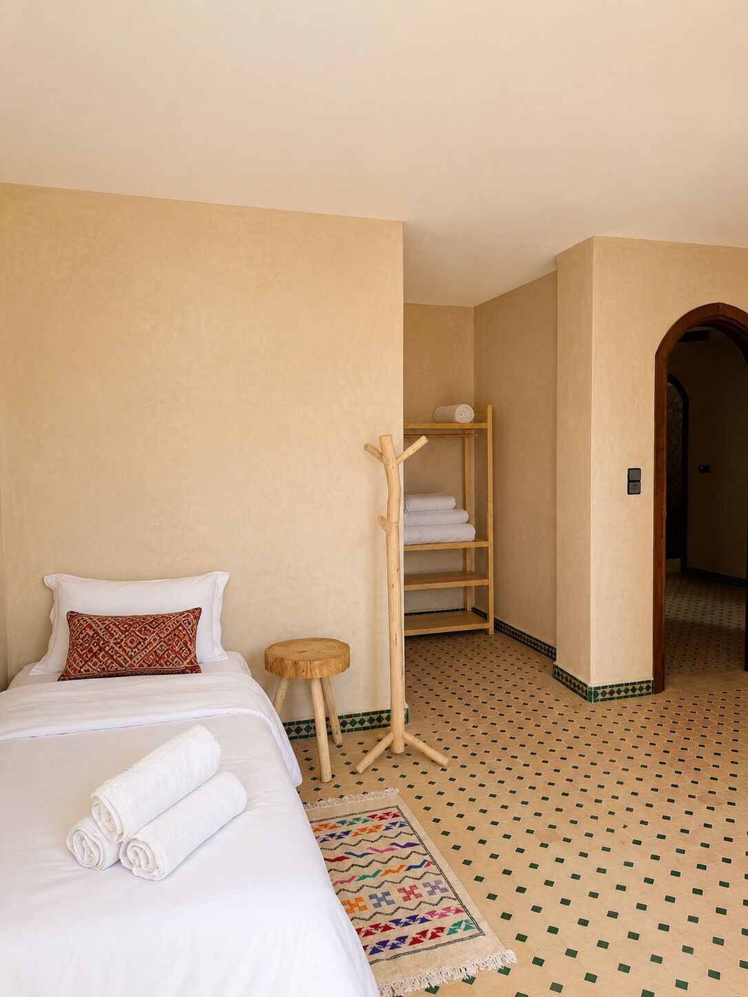 A hotel room with a bed, a wooden side table, a patterned rug, and a wooden shelf with towels.
