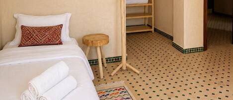 A hotel room with a bed, a wooden side table, a patterned rug, and a wooden shelf with towels.