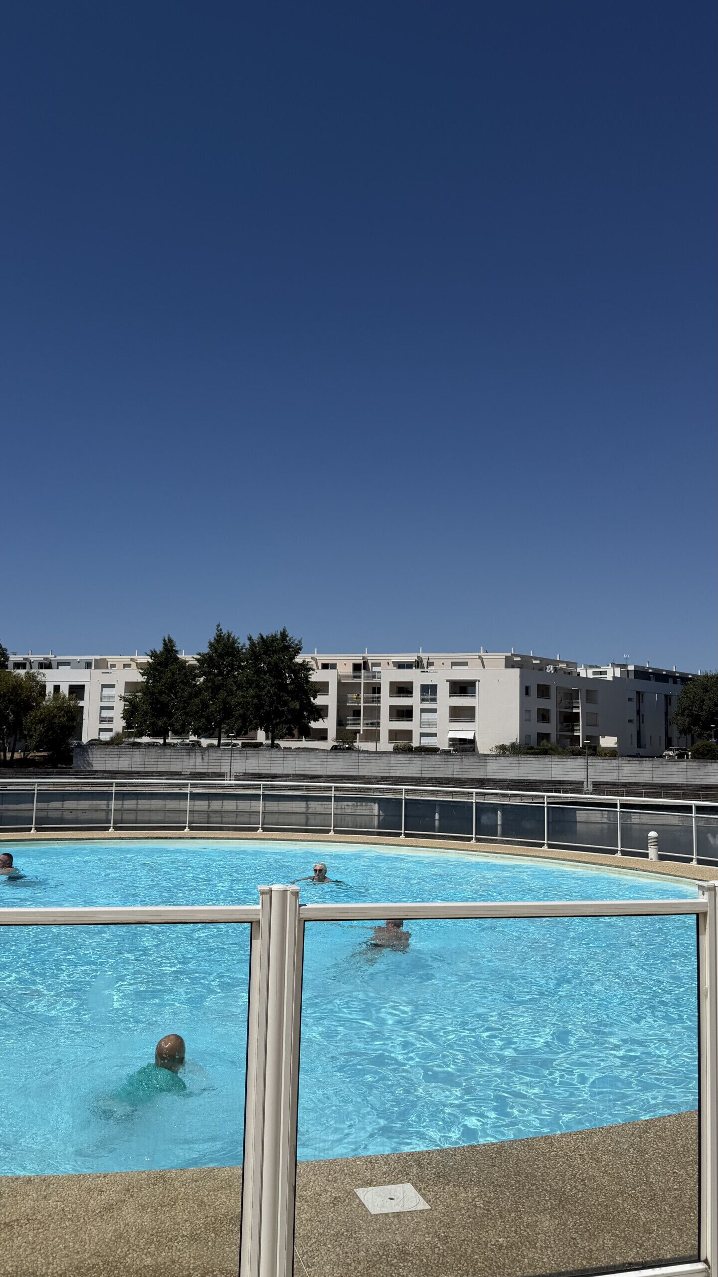 Une piscine à l’eau d’un bleu limpide, entourée d’une clôture. Au fond, on aperçoit des immeubles à plusieurs étages dotés de balcons.