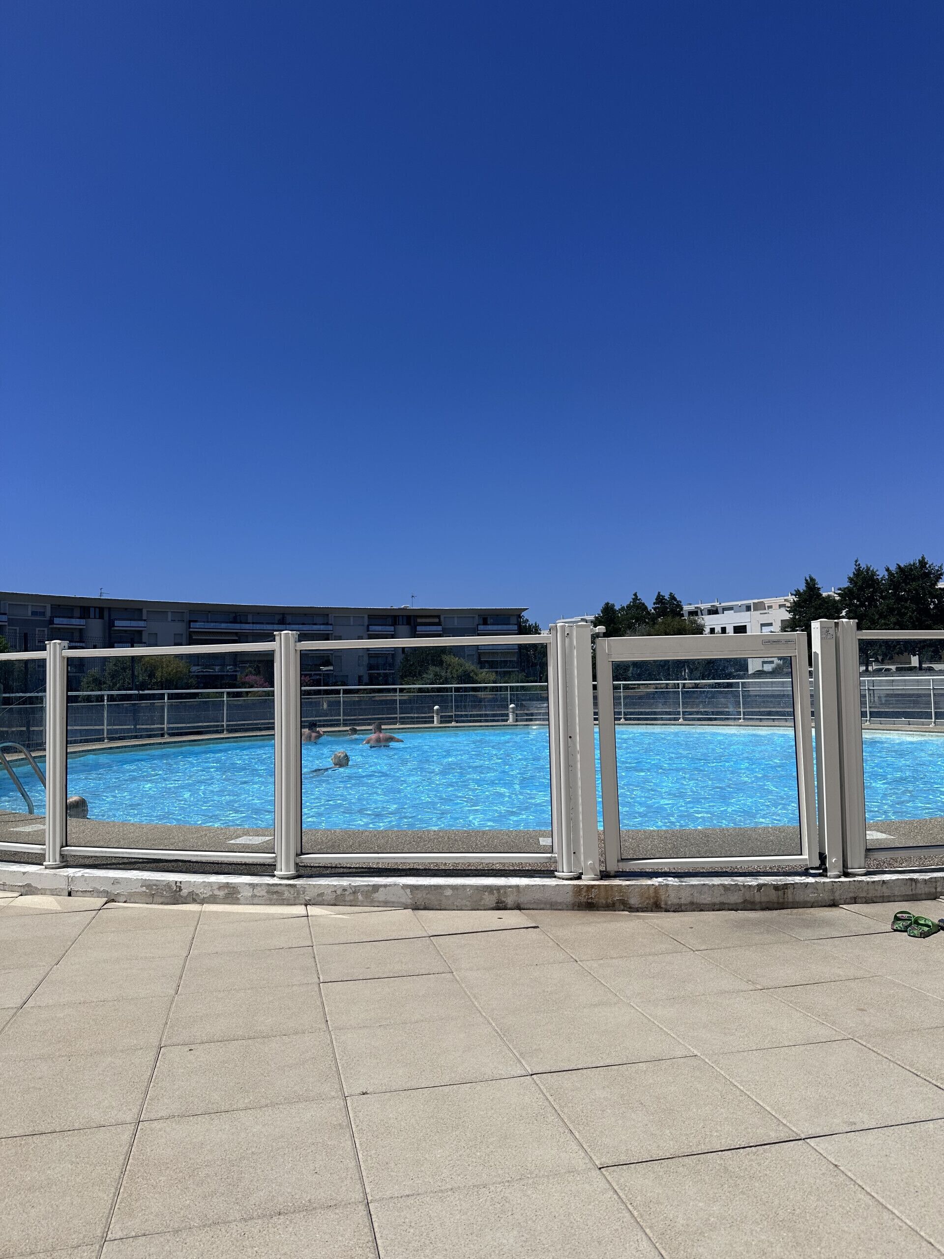 A swimming pool with a clear blue sky above, surrounded by a metal fence and a paved walkway.