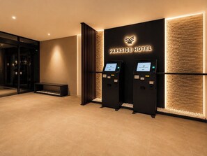 Ein Empfangsbereich mit einer schwarzen Wand, auf der das Logo des Parkside Hotel zu sehen ist, zwei Selbstbedienungs-Check-in-Terminals, einer Sitzbank und einem Glastür-Eingang.