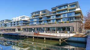A modern multi-story building with a reflective water body in front, featuring balconies and outdoor seating areas. - Sub-Penthouse with Lake Views | Kingston Foreshore (Kingston)