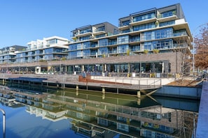 A modern multi-story building with a reflective water body in front, featuring balconies and outdoor seating areas. - Sub-Penthouse with Lake Views | Kingston Foreshore (Kingston)