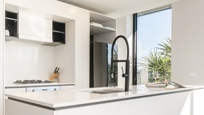 A modern kitchen with white cabinetry, a stainless steel refrigerator, a black faucet, and a window providing a view of palm trees. - Sub-Penthouse with Lake Views | Kingston Foreshore (Kingston)