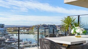 A balcony with a table, chairs, a vase with flowers, and a view of a marina and buildings across a body of water. - Sub-Penthouse with Lake Views | Kingston Foreshore (Kingston)