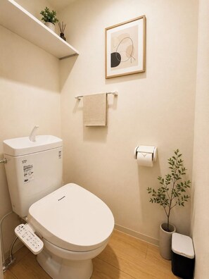 Ein modernes Badezimmer mit Toilette, einem Pflanzenregal, einem gerahmten Kunstwerk, einem Handtuchhalter und einem Papierkorb.