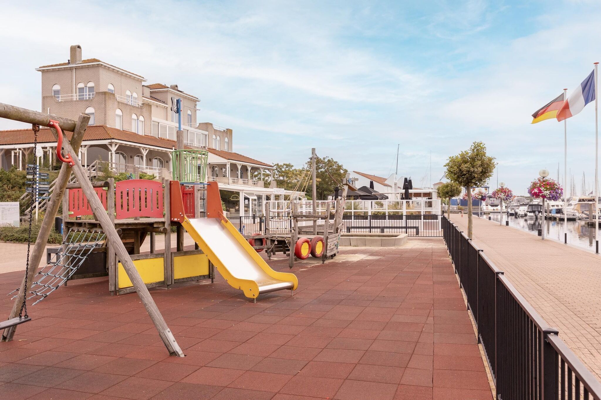 A playground with a slide and climbing structures, a paved walkway, and a building with a balcony in the background.