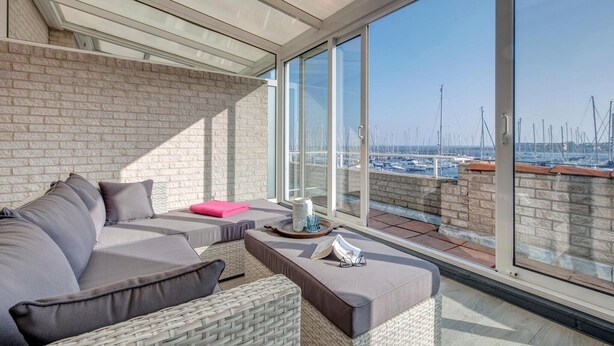 Un balcón con vista a un puerto deportivo, un sofá, una pequeña mesa con un libro y un jarrón, y una silla de mimbre.
