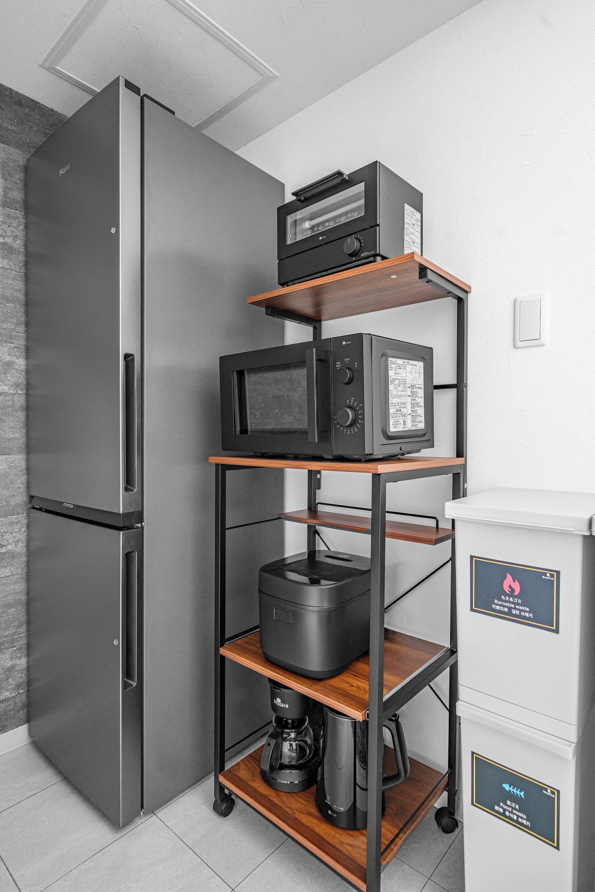 A kitchen with a stainless steel refrigerator, a microwave, a coffee maker, and a shelf unit with appliances.