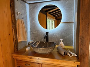 Un baño con un mueble de tocador de madera, un espejo ovalado grande, un lavabo negro y una toalla colgada en un aro metálico.