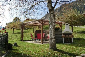 Ein überdachter Außenbereich mit Tischen und Stühlen, eine Holzhütte mit Schornstein und ein Baum im Vordergrund.