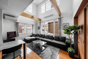 Ein modernes Wohnzimmer mit einem Ecksofa, einem Glastisch, einem Fernseher und einem großen Fenster mit Vorhängen.