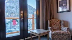 Ein Zimmer mit einem Holztisch, einem gemusterten Sessel, einer Tür mit Blick auf einen verschneiten Berg und einem gerahmten Bild an der Wand.