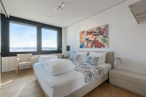 A modern bedroom with a large bed, a chair, a nightstand, a painting, and a view of the city. - Skyflats Vienna South View (Wien)