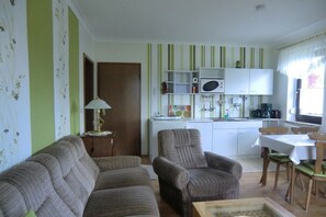 En stue med sofa, lænestol og lille bord. Der er et tekøkken med køleskab, mikroovn og et spisebord med stole.