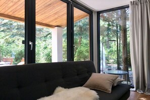 A modern room with a black sofa, a white fluffy pillow, and a view of a forest through large glass windows. - Bungalow in the forest (Borkwalde)