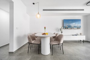A modern dining area with a round table, six chairs, a pendant light, a white console table, and a large framed picture on the wall. - Spacious 4BR Balcony Mamad-RenovatedBldg (Bat Yam)