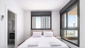 Een moderne hotelkamer met een groot bed, twee ramen, uitzicht op gebouwen en een badkamer met douche.
