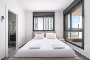 A modern hotel room with a large bed, two windows, a view of buildings, and a bathroom with a shower. - Spacious 4BR Balcony Mamad-RenovatedBldg (Bat Yam)