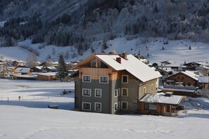 Ein mehrgeschossiges Gebäude mit einem geneigten Dach, umgeben von schneebedeckten Häusern und Bergen.