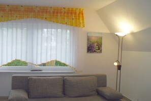Ein Wohnzimmer mit einer Couch, einem Fenster mit Vorhängen, einem Bild an der Wand und einer Stehlampe.