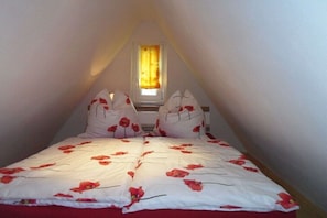 Ein Schlafzimmer mit schrägen Wänden, einem Fenster mit gelben Vorhängen und einem Bett mit einer weiß-rot blumigen Tagesdecke.