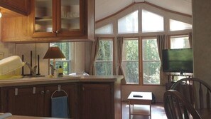 Una cocina con armarios de madera, una ventana con cortinas, una mesita y un televisor.