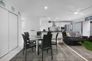 A modern kitchen with white cabinets, a dining area with a glass table and black chairs, and a living area with a sofa and a ceiling fan. - Spacious 2-Bed Stay on Marine Parade (Southport)