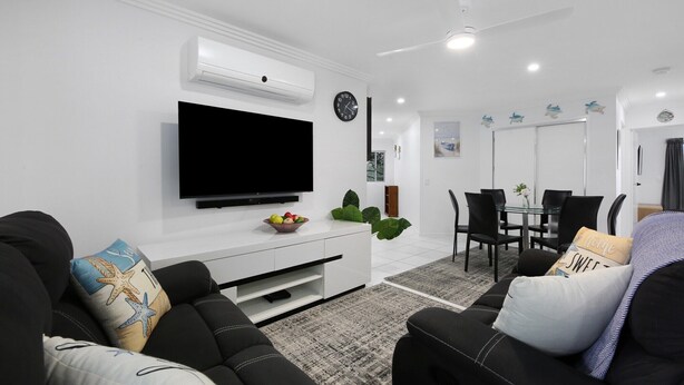 Ett modernt vardagsrum med en platt-TV, svarta soffor, en vit TV-bänk och en matplats med ett glasbord och stolar.