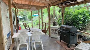 Un patio extérieur abrité, avec une table et des chaises en bois, un barbecue et un petit chariot contenant des ustensiles de cuisine.