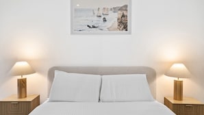 Una cama bien hecha con sábanas blancas, dos mesitas de noche con lámparas, y un cuadro con un paisaje costero en la pared.