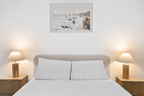 Ein ordentlich bezogenes Bett mit weißen Leinen, zwei Nachttische mit Lampen und ein gerahmtes Bild einer Küstenlandschaft an der Wand.