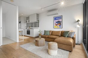 Ein modernes Wohnzimmer mit einer braunen Ledersofa, einem runden Couchtisch und einem gewebten Hocker. Im Hintergrund ist eine Küche mit weißen Schränken und ein Essbereich zu sehen.