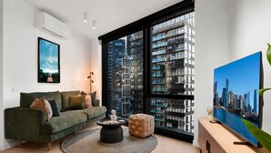 Un salon moderne avec un canapé vert, une table basse ronde, une grande fenêtre donnant sur la ville et un téléviseur à écran plat posé sur une console en bois.