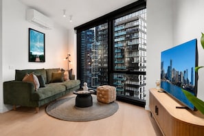 Ein modernes Wohnzimmer mit einem grünen Sofa, einem runden Couchtisch, einem großen Fenster mit Blick auf die Stadt und einem Flachbildfernseher auf einer Holzkonsole.