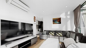 Ein modernes Wohnzimmer mit einem großen Flachbildfernseher, einem grauen Sofa, einem weißen Couchtisch und einer Küche im Hintergrund.