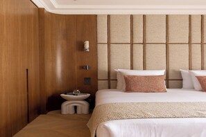 Ein Hotelzimmer mit Holzschrank, Nachttisch mit Fernbedienung, Bett mit weißer Bettwäsche und gemusterten Kissen sowie einem strukturierten Kopfteil.