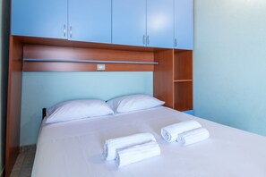 Ein Hotelzimmer mit einem Bett, weißen Leintüchern, zwei aufgerollten Handtüchern und blauen Schränken.