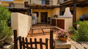 A courtyard with a wooden gate, a stone bench, a potted plant, and a building with a tiled roof and balconies. - Beautiful apartment with sea views (Cannigione)