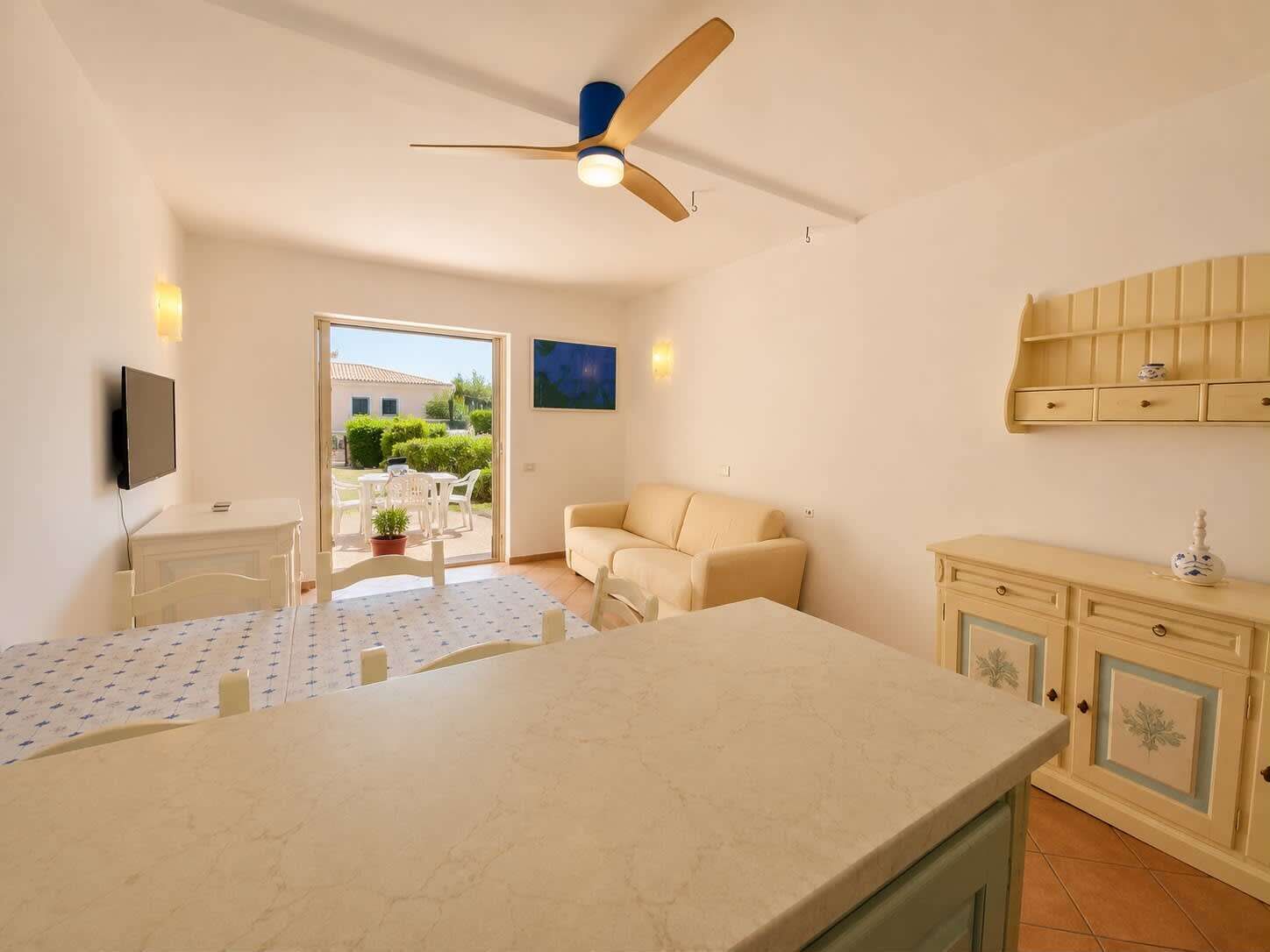 Ein Zimmer mit Deckenventilator, Fernseher, Sofa, Esstisch und einer Küchenzeile mit Schränken.
