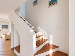 Eine geschwungene Treppe mit Holzstufen und weißen Wänden führt zu einem Raum mit einem beigen Sofa und einem Bild mit Strandmotiv an der Wand.